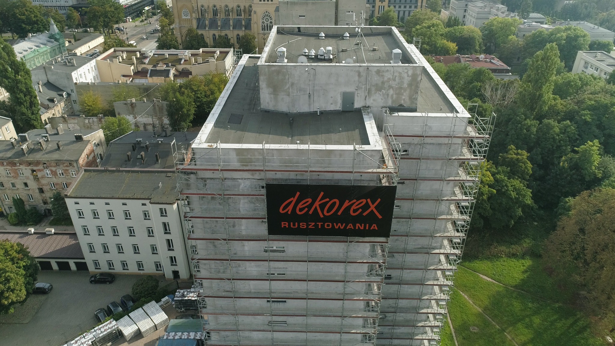 Wysoki budynek w trakcie remontu elewacji, otoczony rusztowaniami i z banerem reklamowym firmy 'dekorex rusztowania', widok z góry na tle zieleni i zabudowy miejskiej.