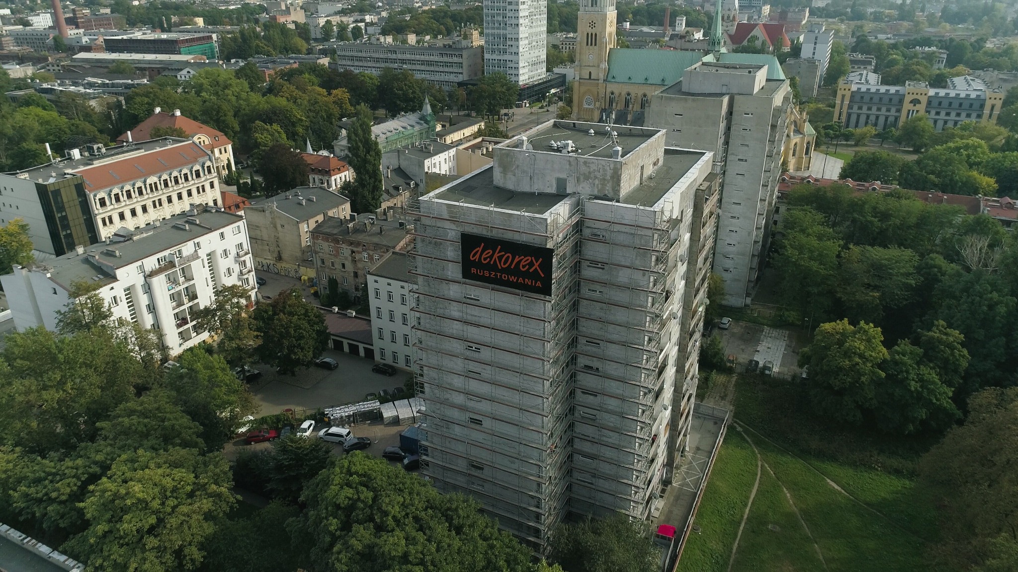 Wysoki budynek mieszkalny w trakcie remontu elewacji, osłonięty rusztowaniami i reklamą firmy rusztowaniowej Dekorex, widok z lotu ptaka na tle zabudowy miejskiej i zieleni.