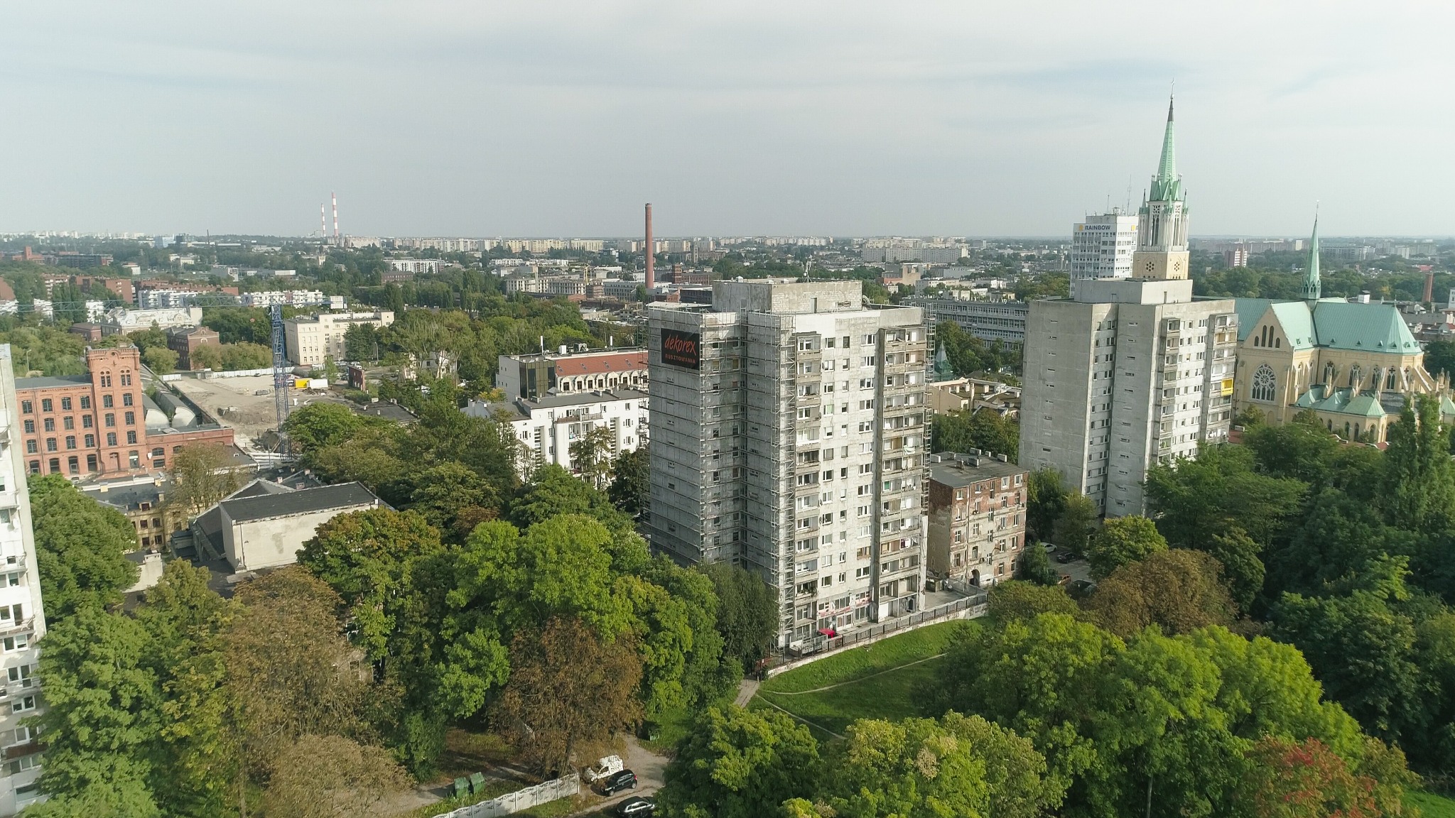 Wysoki budynek mieszkalny w trakcie prac remontowych elewacji, otoczony rusztowaniami i z widocznym banerem reklamowym firmy remontowej, na tle panoramy miasta z kościołem z zielonym dachem.