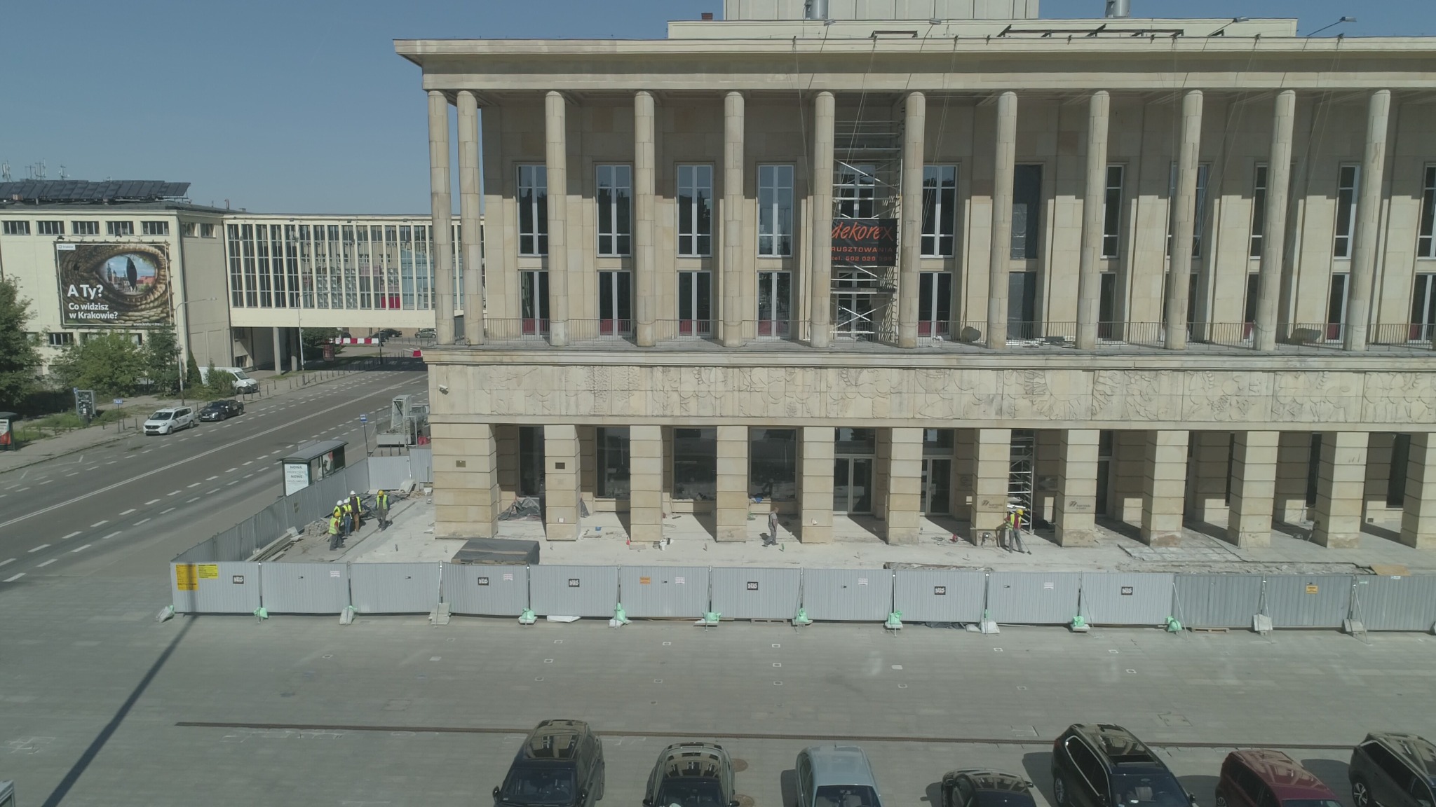 Budynek z kolumnami w trakcie renowacji elewacji, ogrodzony płotem tymczasowym, widoczni pracownicy w kaskach i kamizelkach odblaskowych, rusztowania przy fasadzie.