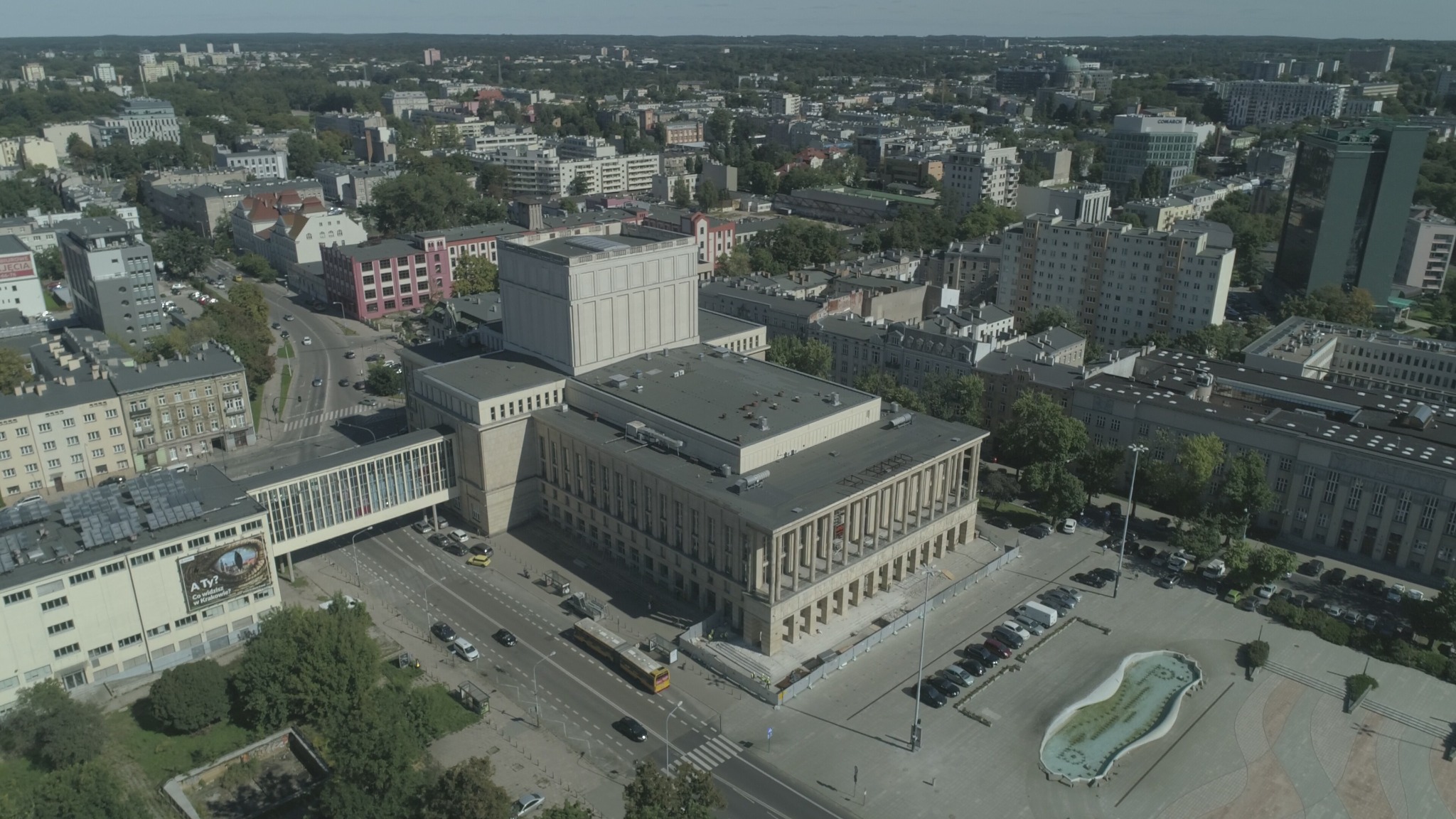 Panorama miejska z lotu ptaka, widoczny budynek teatru z kolumnadą, plac z fontanną, ulice z ruchem samochodowym i tramwajowym, w tle zabudowa miejska i zieleń parków.