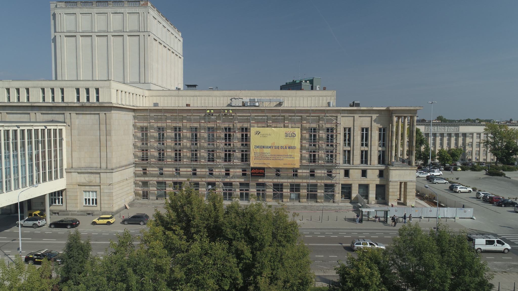 Budynek teatru w rusztowaniach podczas renowacji elewacji, widoczny baner informacyjny z nazwą inwestycji i logotypami firm, pracownicy na rusztowaniu, otoczenie miejskie.