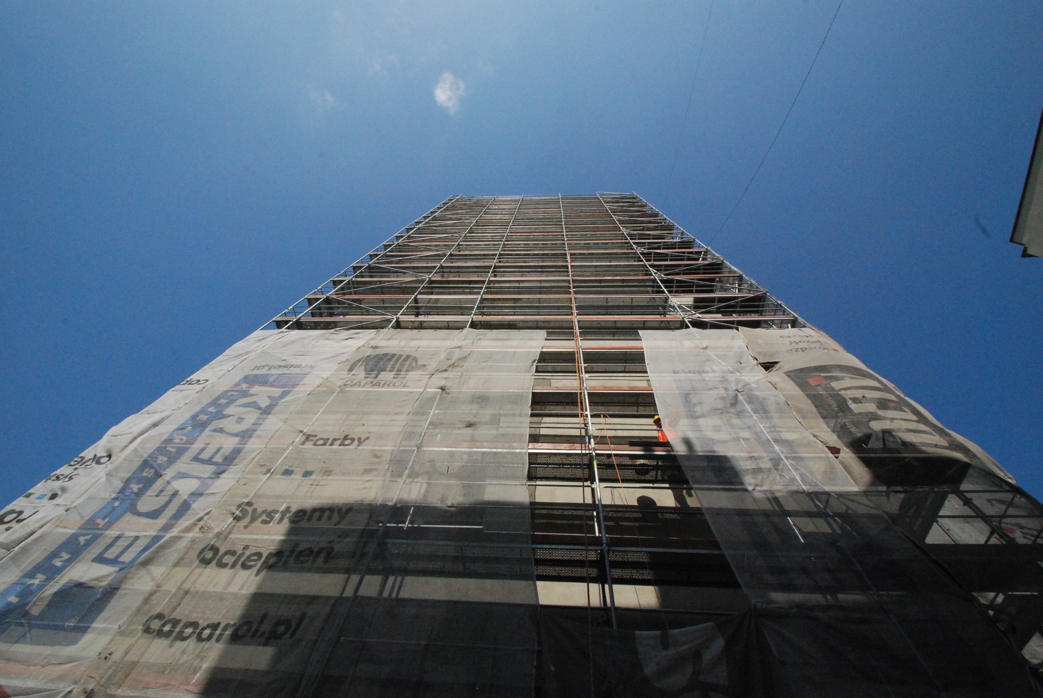 Wysoki budynek w trakcie renowacji, osłonięty siatką z reklamami, widok z dołu na rusztowania i błękitne niebo.