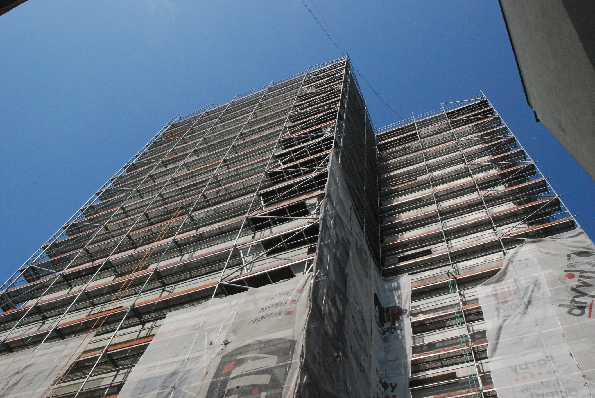 Wysoki budynek w trakcie renowacji, osłonięty rusztowaniami i siatką ochronną, widok z dołu na tle błękitnego nieba.