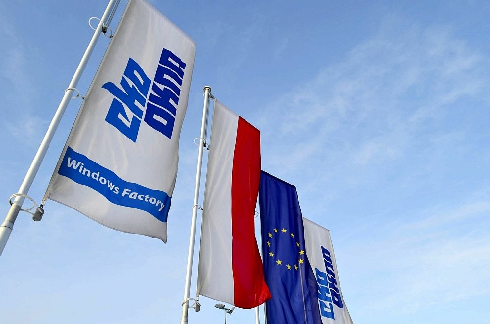 Flagi firmy produkującej stolarkę okienną, w tym flaga z logo, flaga Polski i flaga Unii Europejskiej, na tle błękitnego nieba.