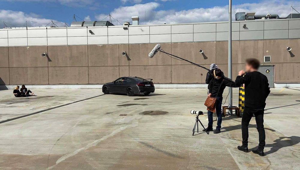Ekipa filmowa kręci ujęcie samochodu na parkingu; mikrofon kierunkowy, kamera na statywie, dwie osoby siedzące w tle.