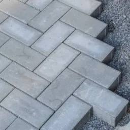 Ułożona szara kostka brukowa w jodełkę sąsiaduje z warstwą podsypki żwirowej i fragmentem betonowej wylewki.