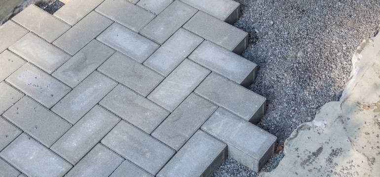 Ułożona szara kostka brukowa w jodełkę sąsiaduje z warstwą podsypki żwirowej i fragmentem betonowej wylewki.