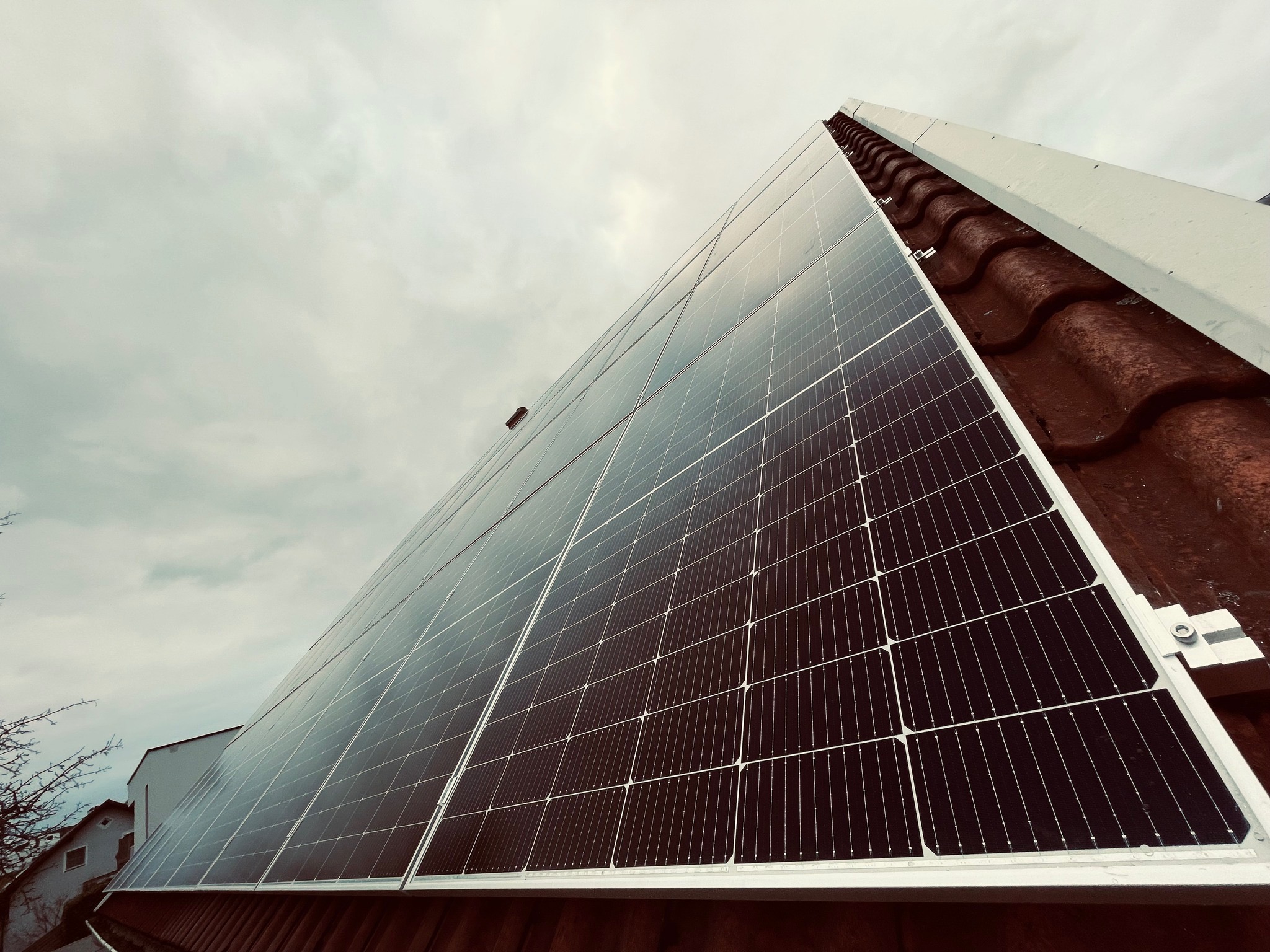Panel słoneczny na dachu z dachówką ceramiczną, widok z dołu pod kątem, pochmurne niebo w tle, z widocznymi elementami mocującymi i fragmentem budynku obok.