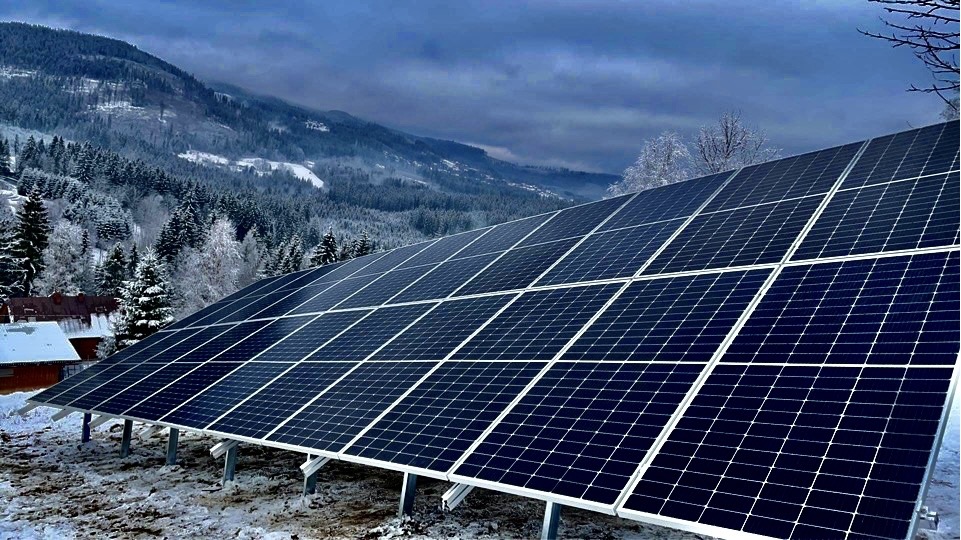 Panel słoneczny w zimowym krajobrazie górskim, widoczne zaśnieżone drzewa i szczyty w tle.