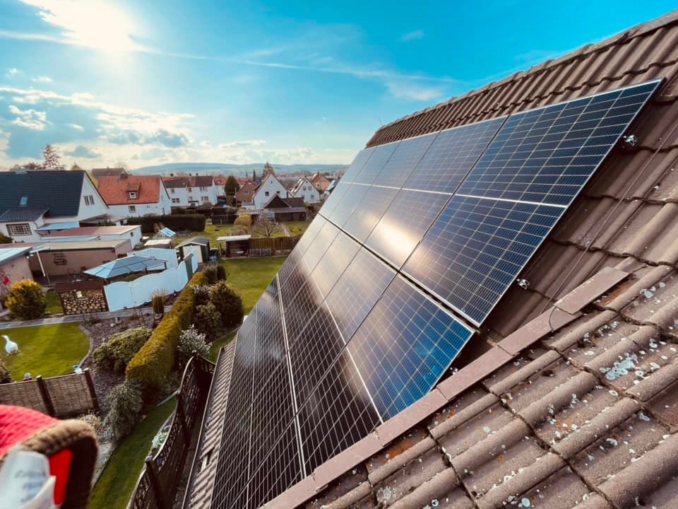 Panele słoneczne zamontowane na brązowym, lekko zabrudzonym mchem dachu domu jednorodzinnego, widok z boku, w tle zabudowa mieszkalna i błękitne niebo.