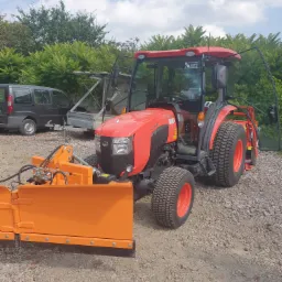 Czerwony traktor Kubota z pomarańczowym pługiem do śniegu stoi na żwirowym podłożu, z otwartymi drzwiami kabiny, widoczny samoch&oacute;d dostawczy i przyczepa w tle.
