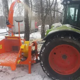 Pomarańczowa rębarka do gałęzi podłączona do traktora Claas na ośnieżonym terenie w trakcie pracy, z widocznymi wylatującymi wi&oacute;rami i drzewami w tle.