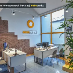 Jasne biuro firmy instalacyjnej z logo 'ekopunkt' na ścianie, biurkami, komputerami i dekoracyjną ścianą z cegły oraz dużym oknem