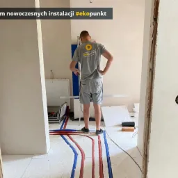 Instalacja ogrzewania podłogowego z rurami w kolorze niebieskim i czerwonym, widok zza pleców instalatora w szarej koszulce z logo firmy, częściowo wykończone ściany w jasnym odcieniu, widoczne...