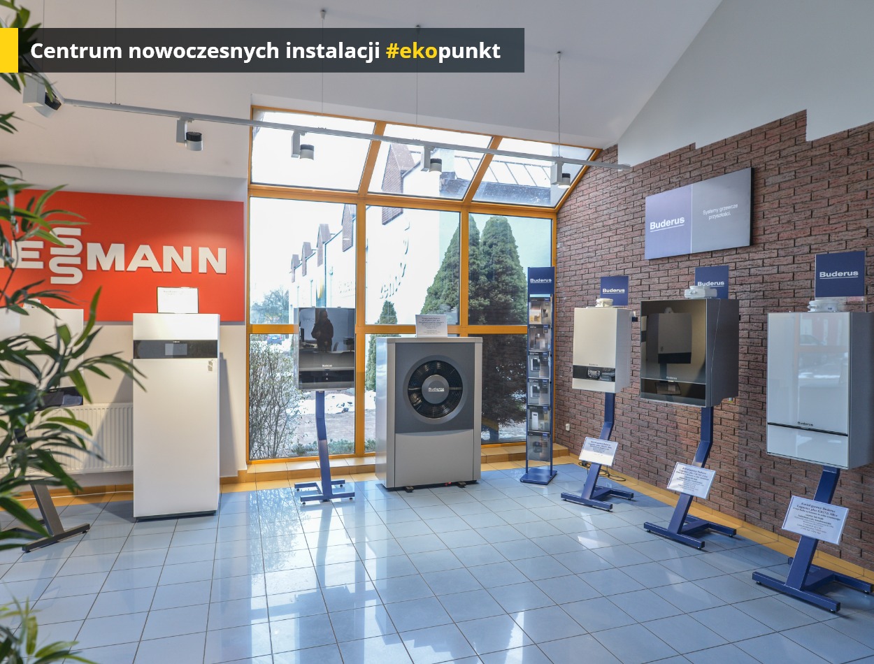 Salon ekspozycyjny z różnymi modelami pomp ciepła marek Viessmann i Buderus, ustawionymi na niebieskich stojakach, z widoczną ścianą z cegły i dużym oknem w tle.