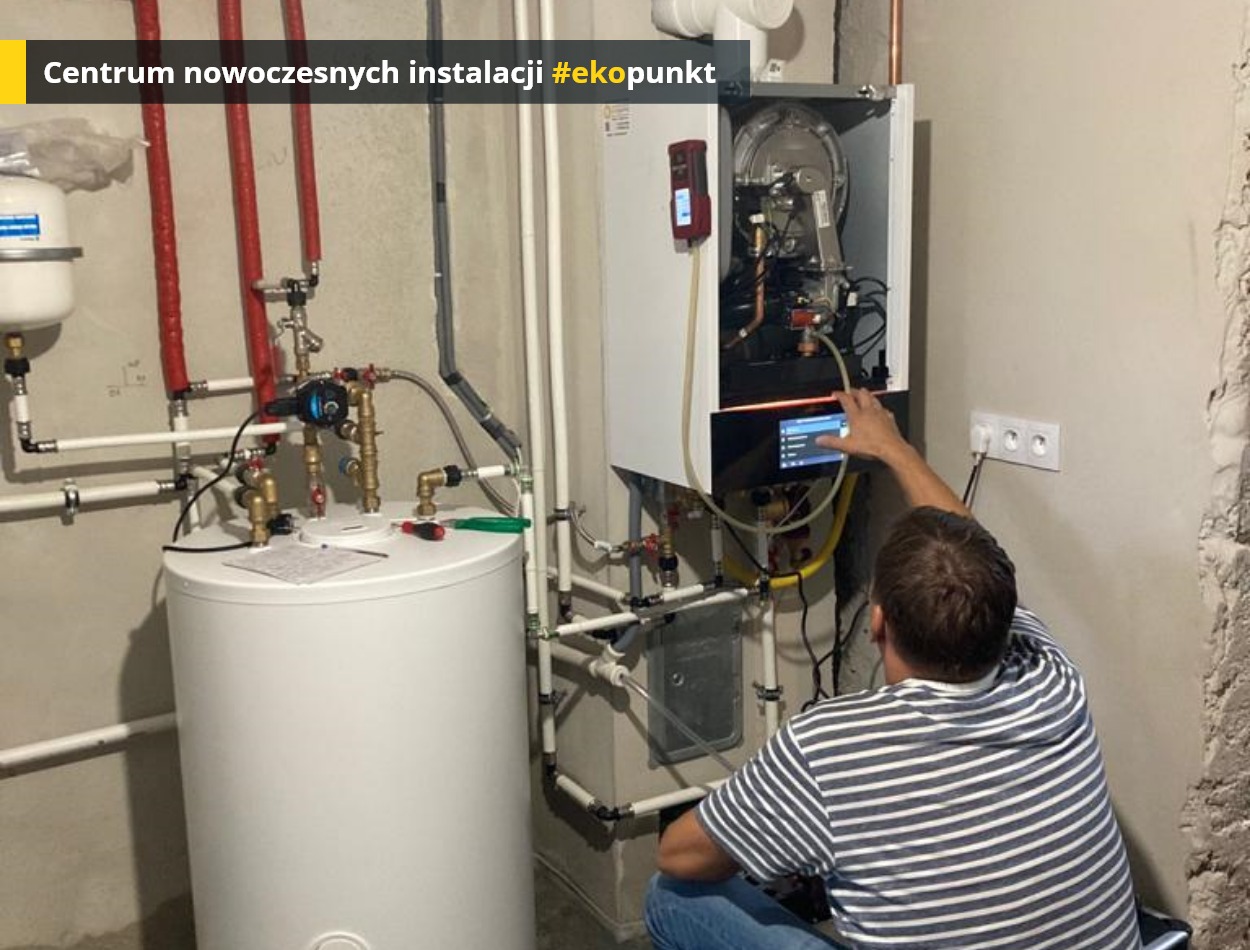 Instalator w paski reguluje ustawienia nowoczesnego pieca grzewczego z dotykowym panelem sterowania, widoczny biały zasobnik wody i rury instalacyjne.