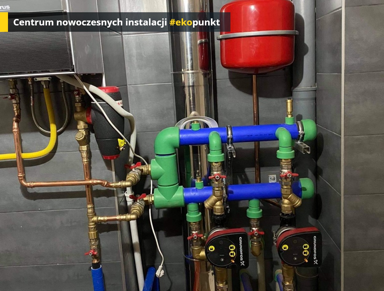 Złożona instalacja hydrauliczna z rur miedzianych, niebieskich i zielonych, wyposażona w dwie pompy obiegowe Grundfos X, zbiornik wyrównawczy w kolorze czerwonym oraz filtr magnetyczny na tle...