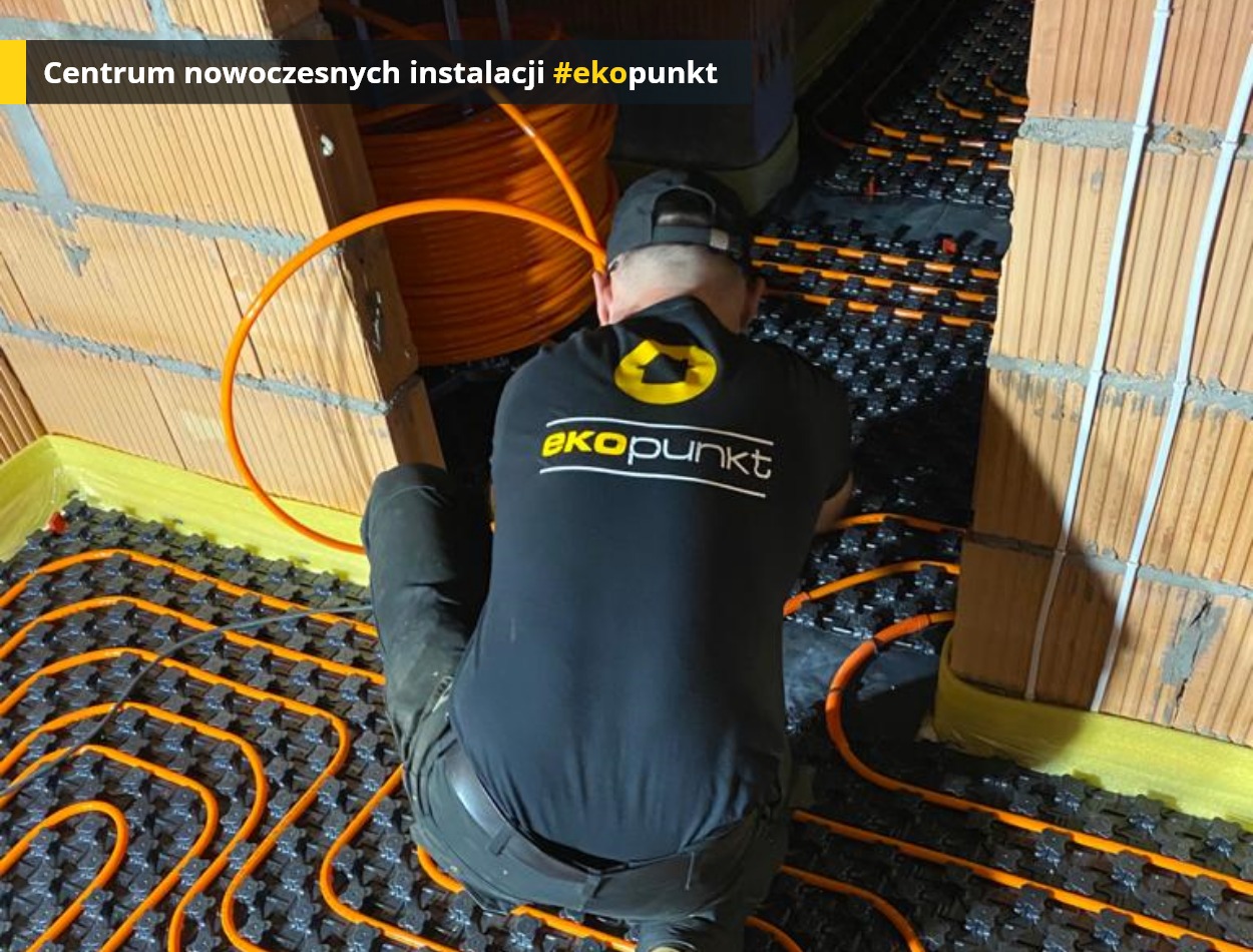 Instalacja ogrzewania podłogowego: mężczyzna w czarnej koszulce z logo 'ekopunkt' układa pomarańczowe rury grzewcze na czarnej macie izolacyjnej w nowo budowanym pomieszczeniu z ceglanymi ścianami,...