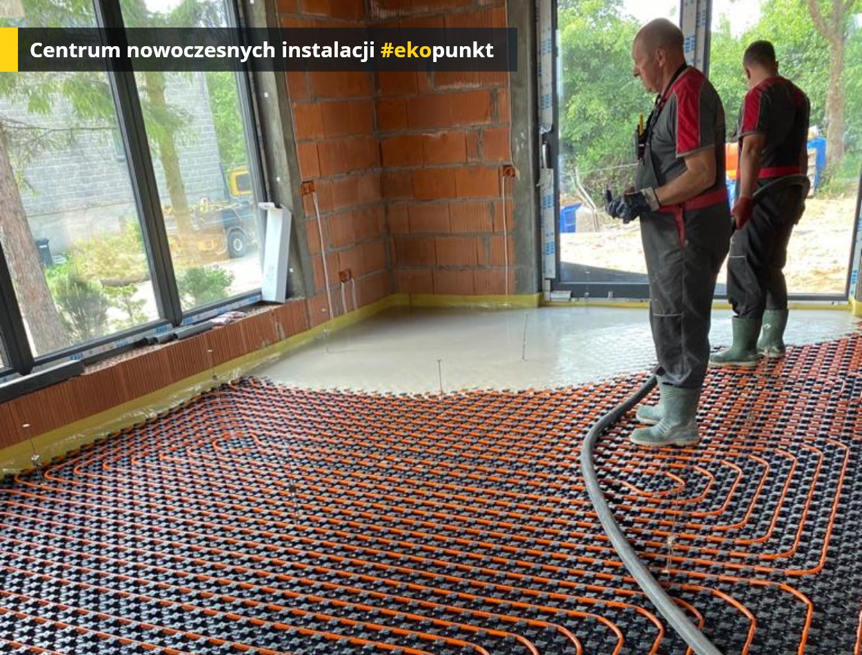 Instalacja ogrzewania podłogowego z wylewką samopoziomującą w surowym wnętrzu z oknami panoramicznymi, widoczni dwaj pracownicy w kombinezonach i gumowych butach podczas pracy.