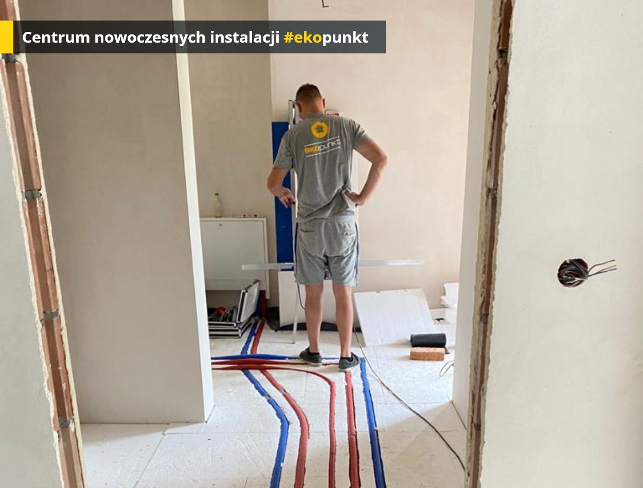 Instalacja ogrzewania podłogowego z rurami w kolorze niebieskim i czerwonym, widok zza pleców instalatora w szarej koszulce z logo firmy, częściowo wykończone ściany w jasnym odcieniu, widoczne...