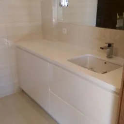 Biała szafka łazienkowa z umywalką i chromowaną baterią, podłużne płytki na ścianie, lustro z oświetleniem w tle.