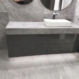 Szara łazienka z szafką pod umywalką z błyszczącymi, grafitowymi frontami, umywalką nablatową i armaturą w kolorze czarnym, dopasowaną do szarej ściany z płytek imitujących kamień.