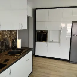 Nowoczesna kuchnia z białymi, lakierowanymi frontami, czarnym blatem imitującym marmur, oraz zabudowaną lodówką i piekarnikiem; na blacie leżą okulary i telefon.