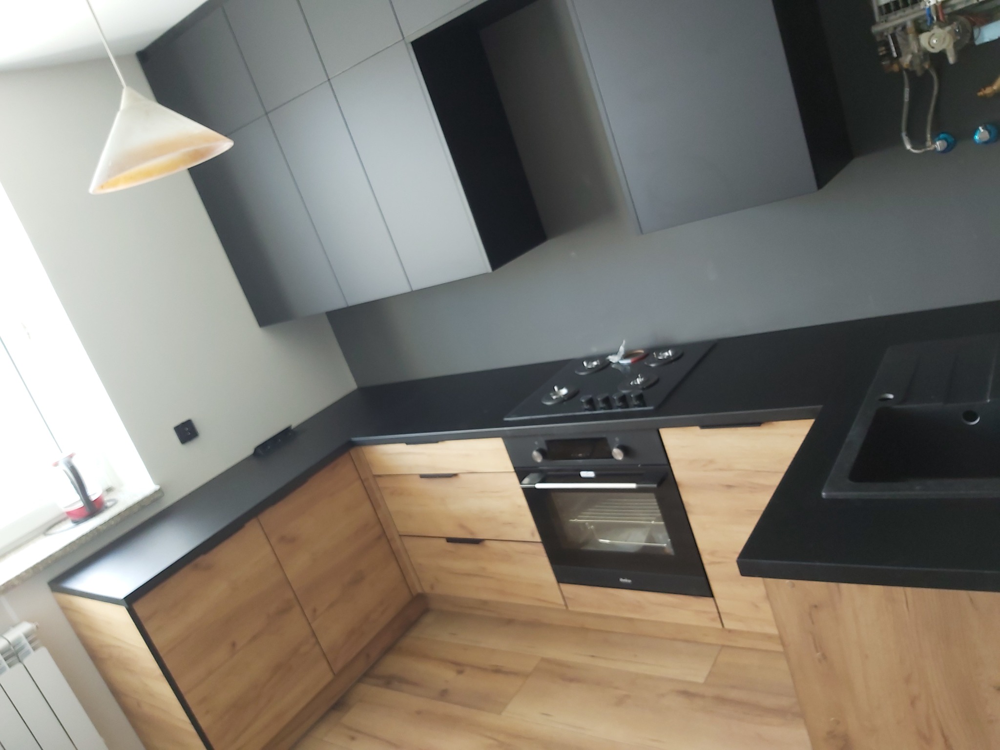 Nowoczesna kuchnia z drewnopodobnymi frontami szafek dolnych i grafitowymi szafkami wiszącymi, czarny blat, piekarnik w zabudowie i płyta gazowa, widoczny fragment okna i lampy.