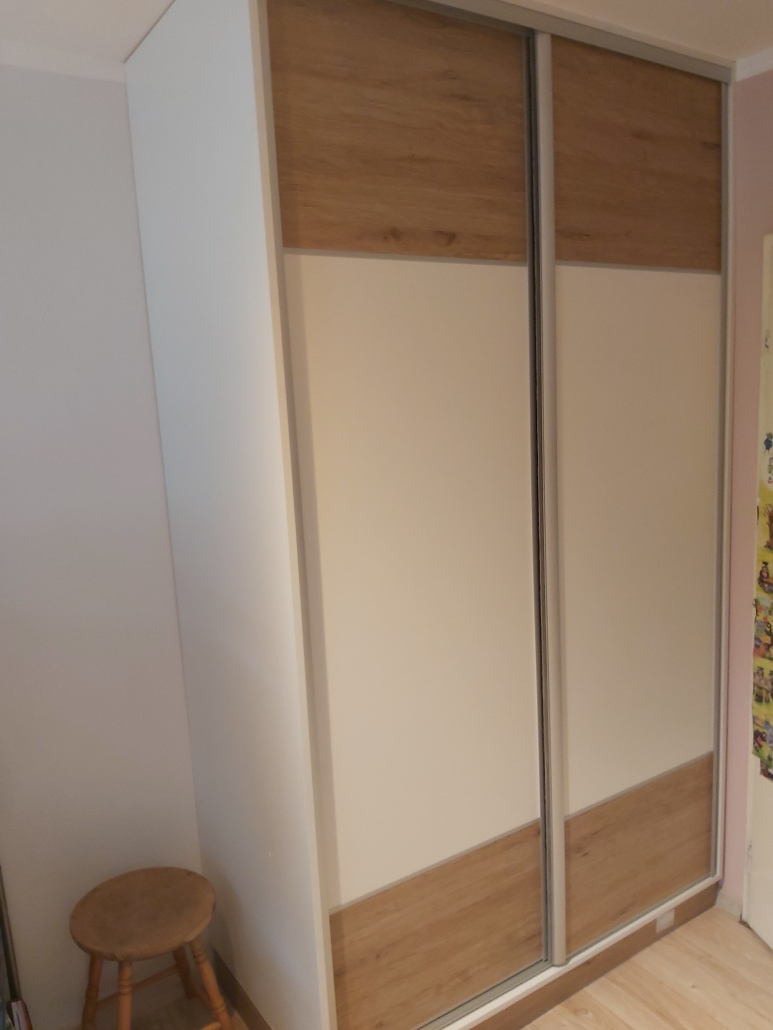 Dwudrzwiowa szafa przesuwna z białymi panelami i drewnopodobnymi akcentami na górze i dole, widoczny drewniany taboret z przodu.