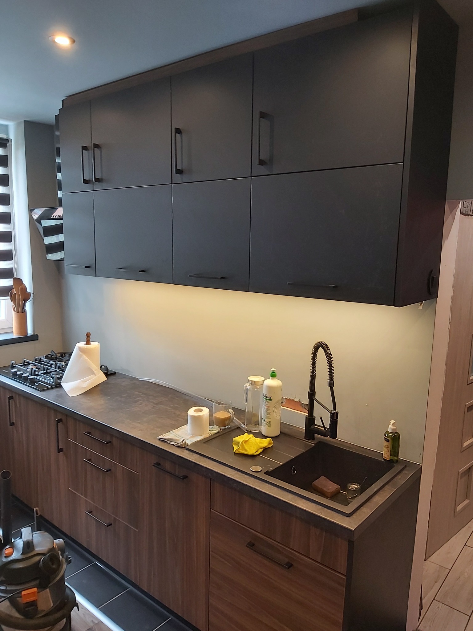 Nowoczesna kuchnia z ciemnymi, matowymi szafkami górnymi i dolnymi z drewnopodobnym frontem, czarnym zlewem i baterią, oświetleniem LED pod szafkami, oraz częściowo widocznym odkurzaczem...