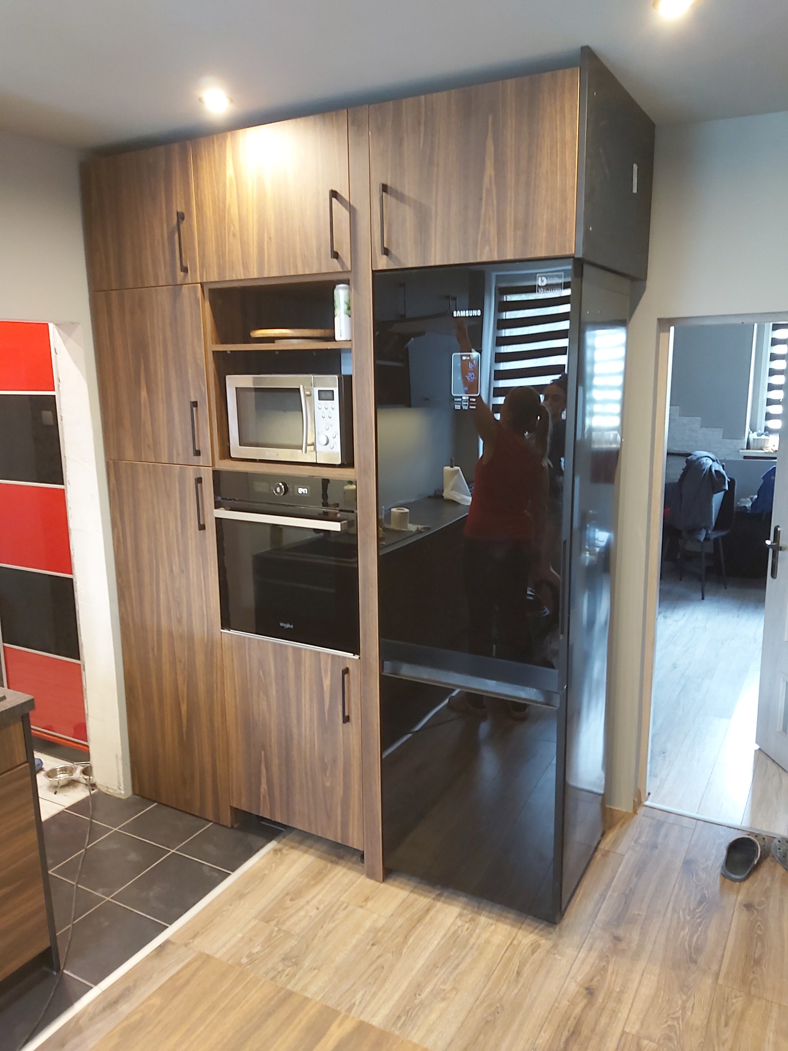 Wysoka zabudowa kuchenna z forniru w odcieniach brązu, zawierająca piekarnik, mikrofalówkę i lodówkę Samsung, widoczna granica płytek podłogowych i paneli.