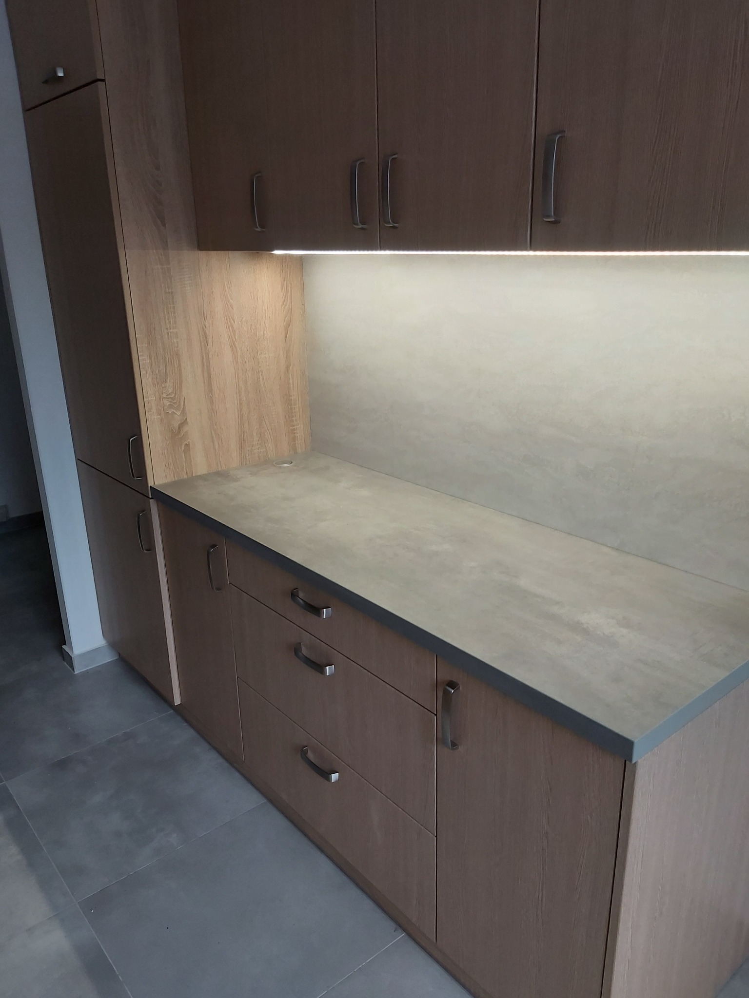 Jasne drewniane szafki kuchenne z szarym blatem i podświetleniem LED pod górnymi szafkami, widoczne chromowane uchwyty.