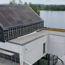 Nowoczesny budynek z aluminiowymi oknami i dachem pokrytym panelami słonecznymi, widok z góry na jezioro w tle, widoczne rusztowania wskazujące na trwające prace wykończeniowe.