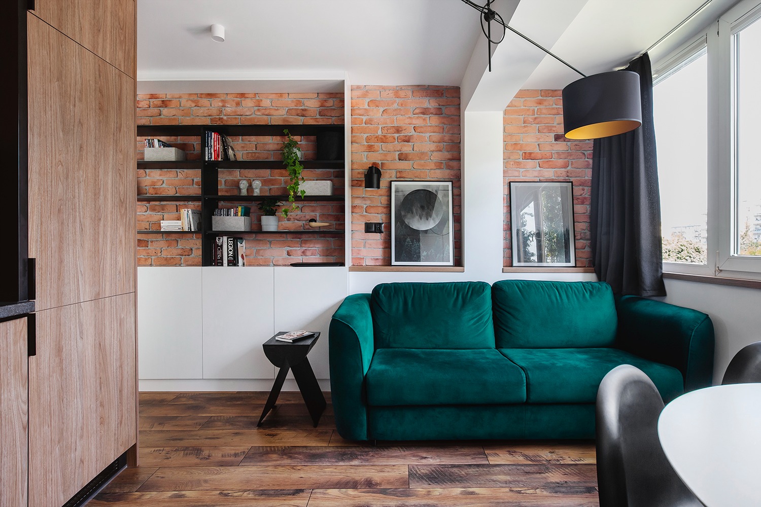 Salon z ceglaną ścianą, zieloną sofą i drewnianą podłogą. Na ścianie półki z książkami i dekoracjami, a obok okna wisząca lampa z czarnym abażurem.