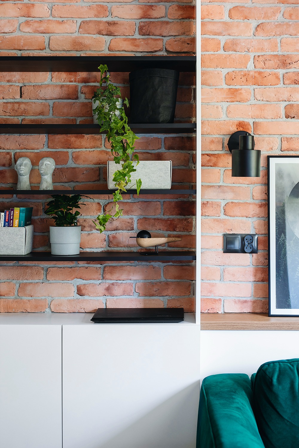 Regał z czarnymi półkami na tle ceglanej ściany, ozdobiony roślinami, figurkami i drewnianym ptakiem, z białymi szafkami na dole i fragmentem zielonej sofy.