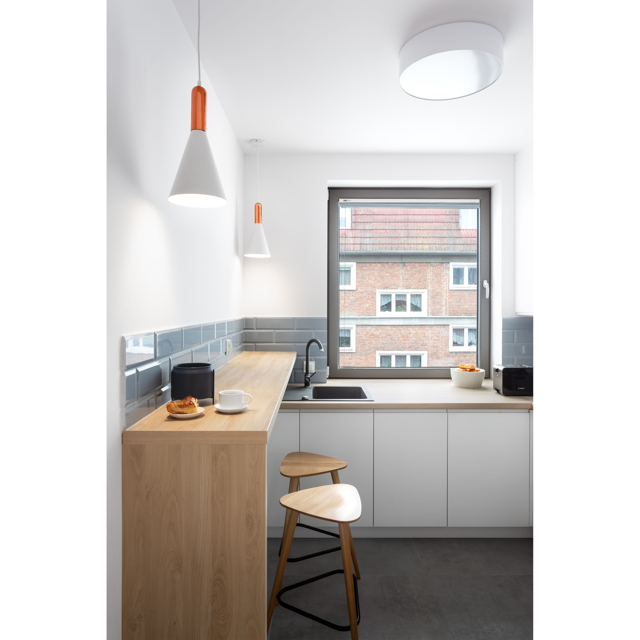Nowoczesna kuchnia z drewnianym blatem, szarymi płytkami i białymi szafkami, oświetlona wiszącymi lampami z miedzianymi akcentami, widok z okna na budynek mieszkalny, na blacie filiżanka i rogalik.