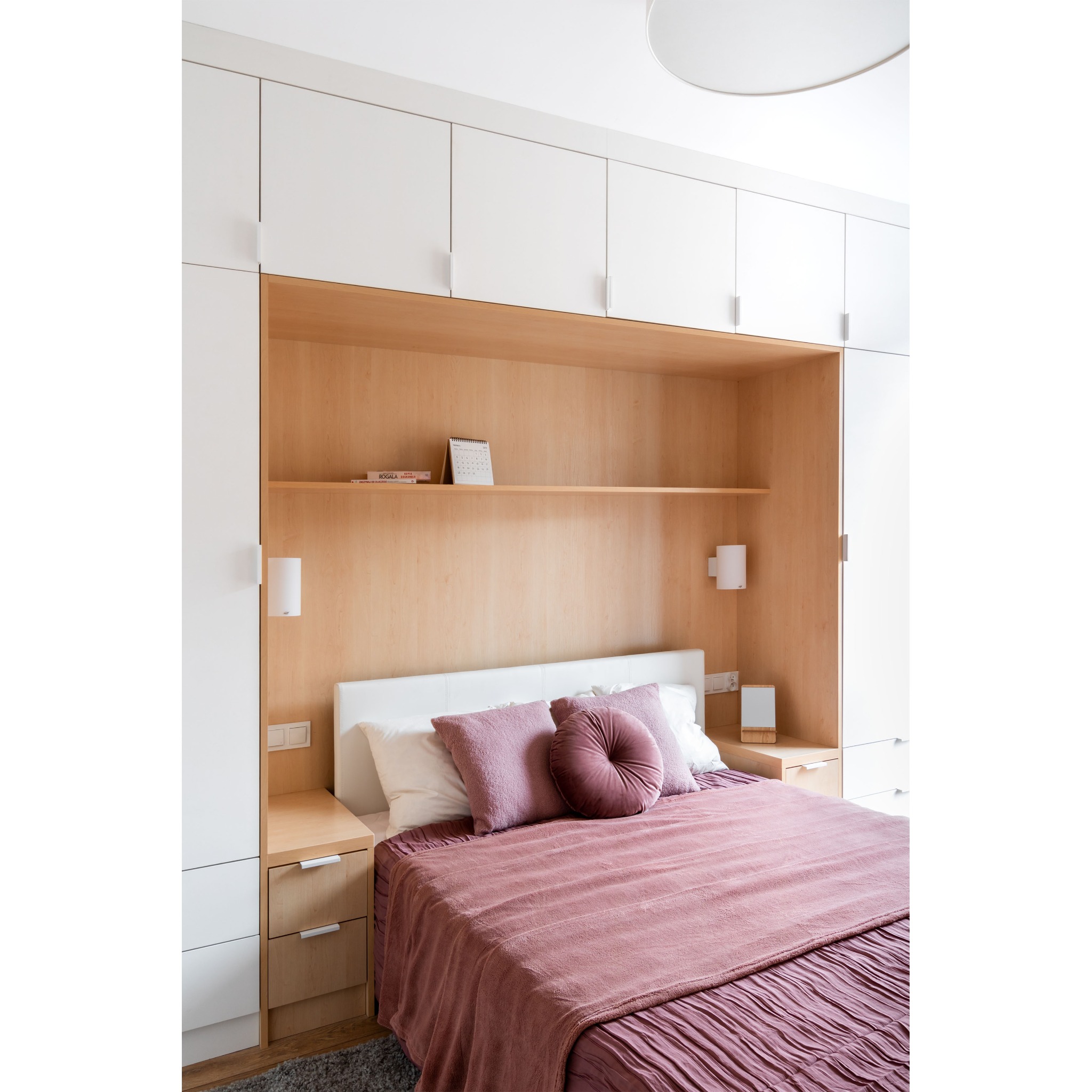 Nowoczesna sypialnia z zabudową meblową z jasnego drewna, białymi szafkami i łóżkiem z różową narzutą oraz okrągłą poduszką.