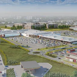 Szeroki plan kompleksu handlowego z parkingiem wypełnionym samochodami, otoczonego zielenią i budynkami mieszkalnymi w tle, widzianego z lotu ptaka.