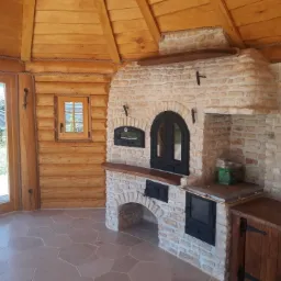Wnętrze drewnianej altany z piecem chlebowym z kamienia, widoczne okno i drzwi przesuwne z drewna, podłoga wyłożona płytkami o nieregularnym kształcie.