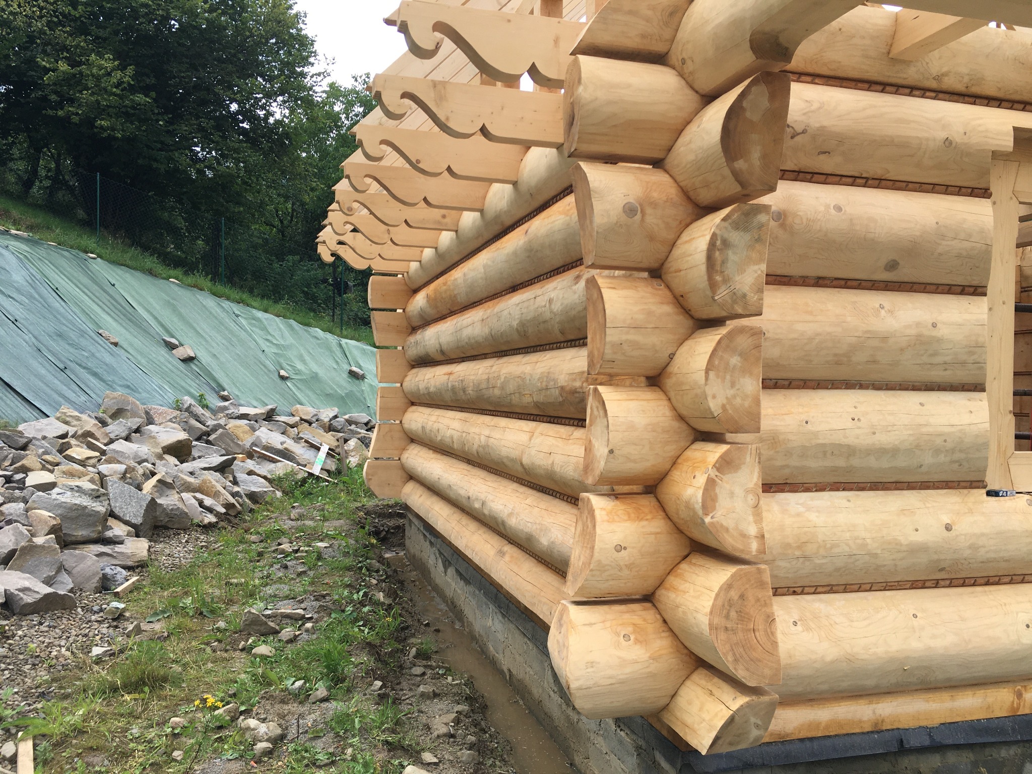 Fragment budowy domu z okrąglaków, widoczne połączenia bali, dekoracyjne zakończenia krokwi, w tle zabezpieczona skarpa.