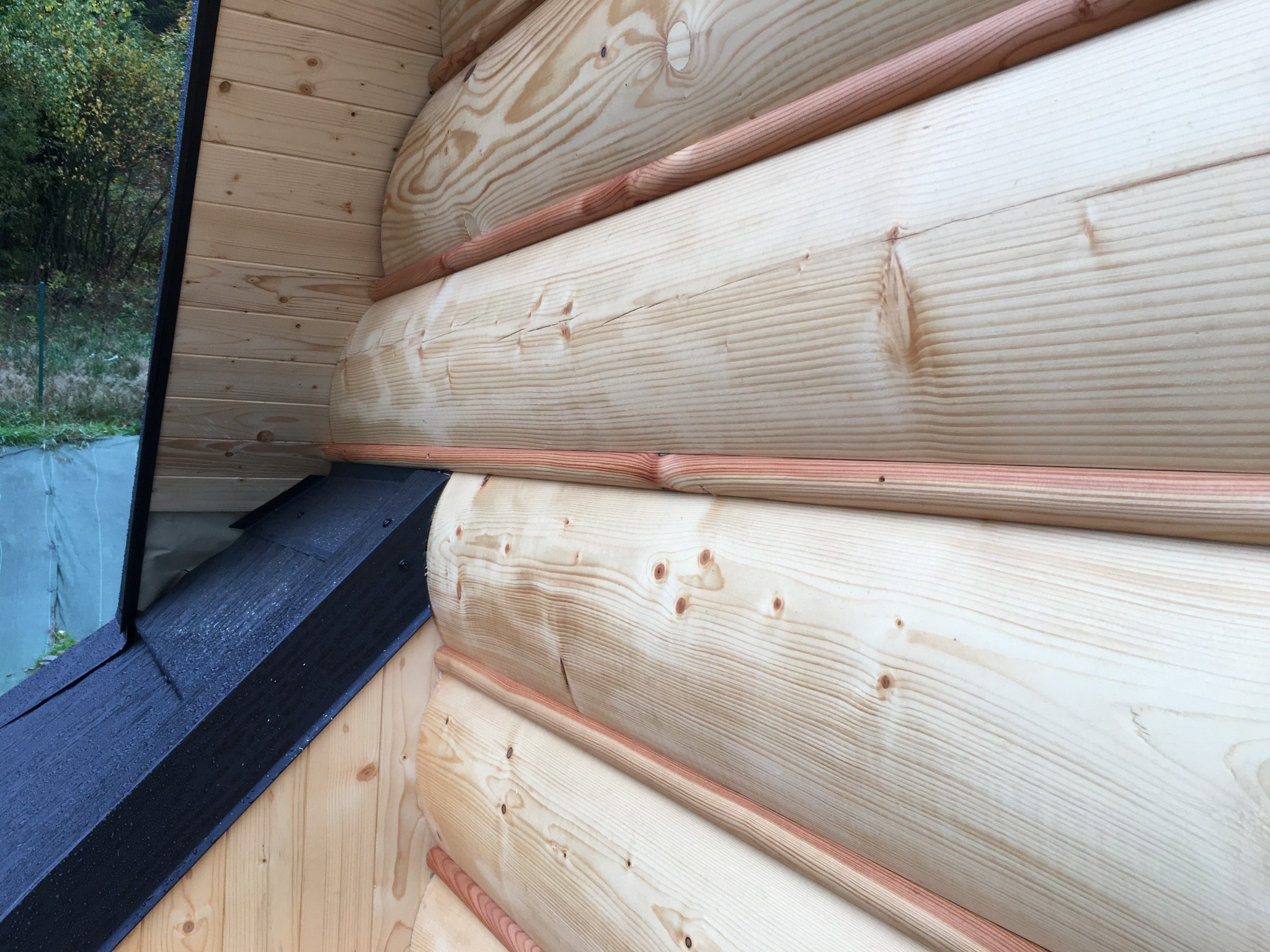 Fragment zewnętrznej ściany domu z bali, z widocznym czarnym elementem dachu i naturalnym rysunkiem drewna.