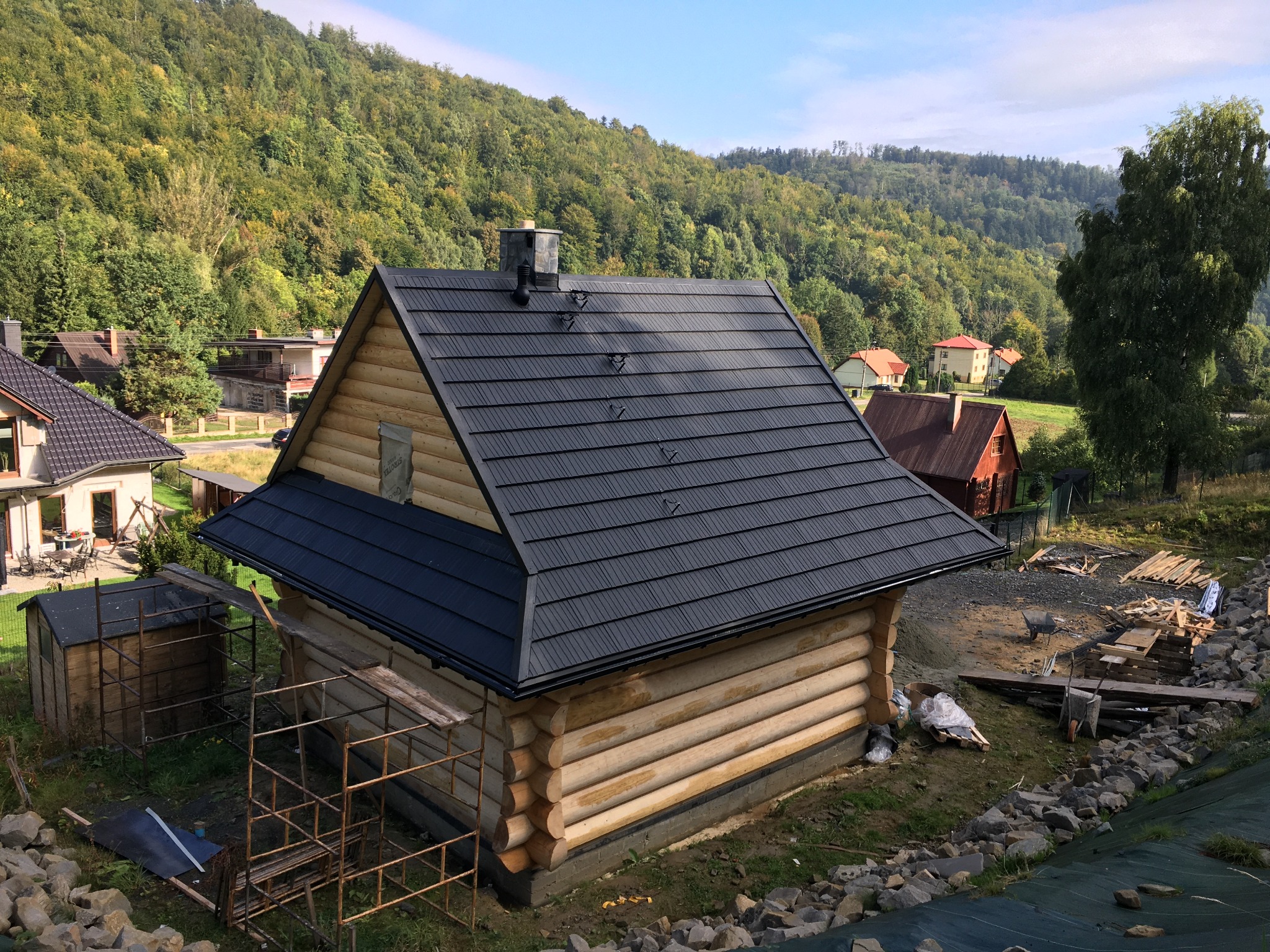Wznoszony dom z bali z czarnym, blaszanym dachem na tle zalesionych wzgórz; widoczne rusztowania i materiały budowlane.