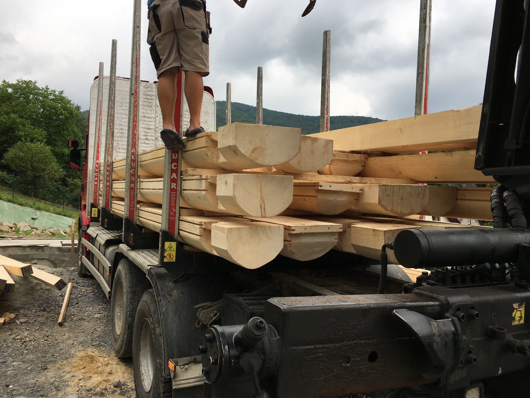 Ciężarówka załadowana elementami konstrukcyjnymi z drewna, widoczne nogi pracownika na górze ładunku, w tle góry i pochmurne niebo.