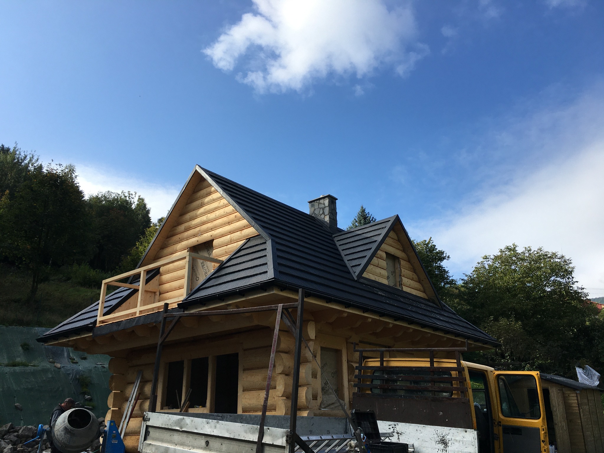 Surowy, drewniany dom w trakcie budowy, z czarnym dachem, widoczne rusztowania i ciężarówka w tle, błękitne niebo z chmurami.