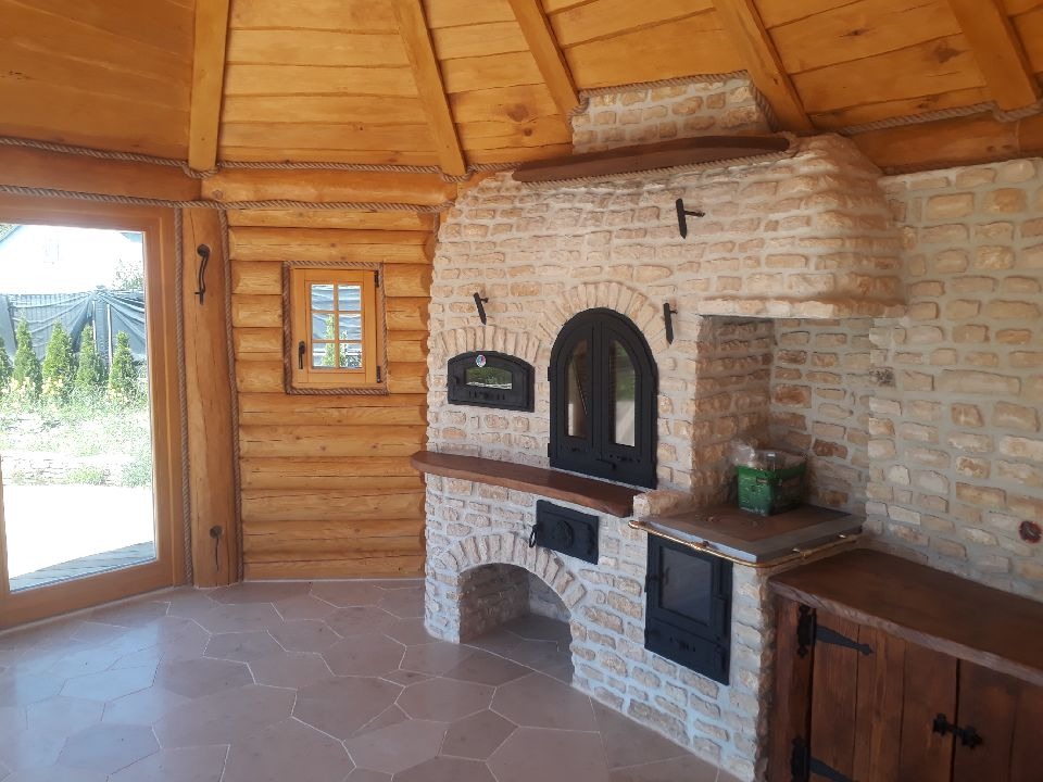Wnętrze drewnianej altany z piecem chlebowym z kamienia, widoczne okno i drzwi przesuwne z drewna, podłoga wyłożona płytkami o nieregularnym kształcie.