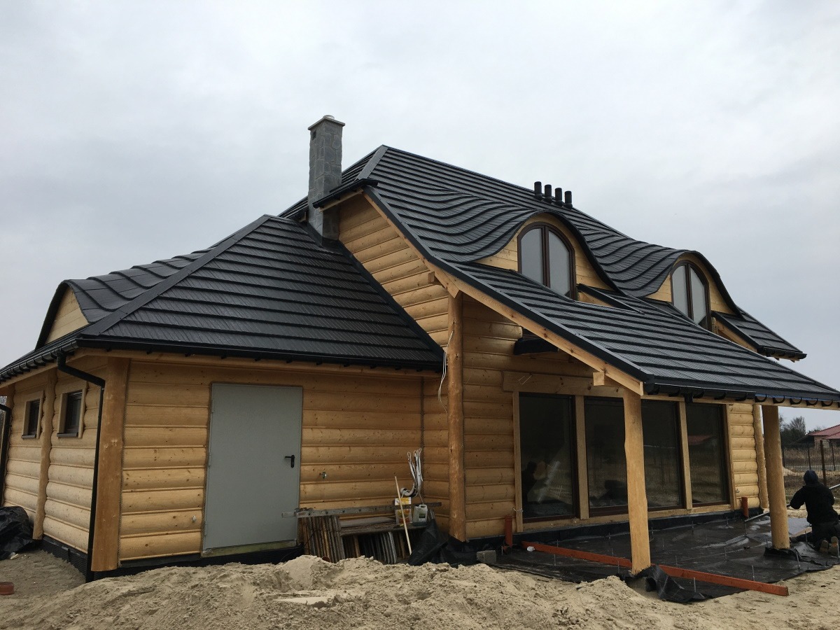 Surowy dom z bali z ciemną, falistą blachodachówką, widoczne okna w lukarnach i komin z kamienia, teren wokół domu częściowo zasypany piaskiem.