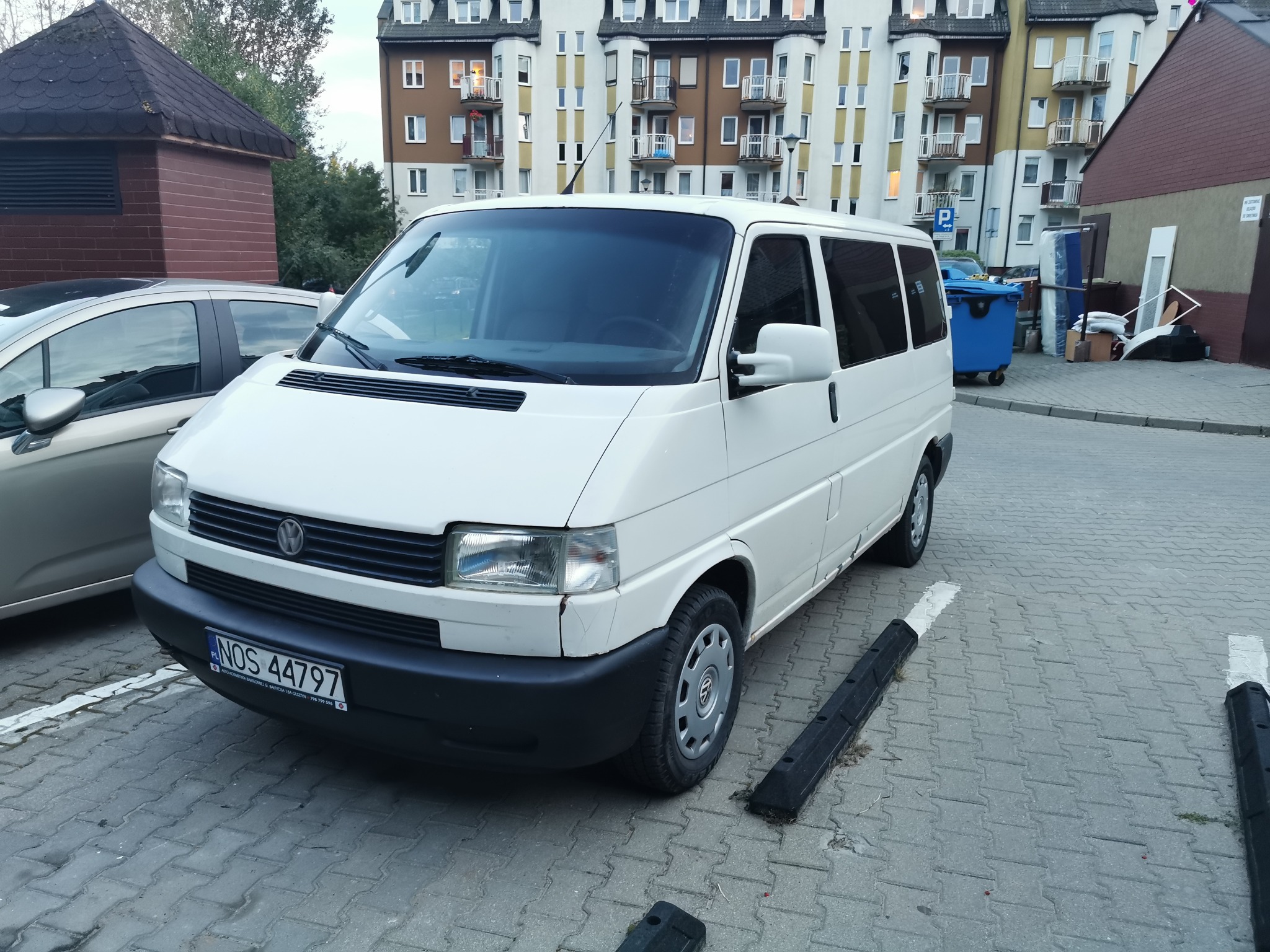 Biały bus Volkswagen Transporter zaparkowany na parkingu z kostki brukowej przed budynkiem mieszkalnym, widoczne ślady użytkowania na karoserii.