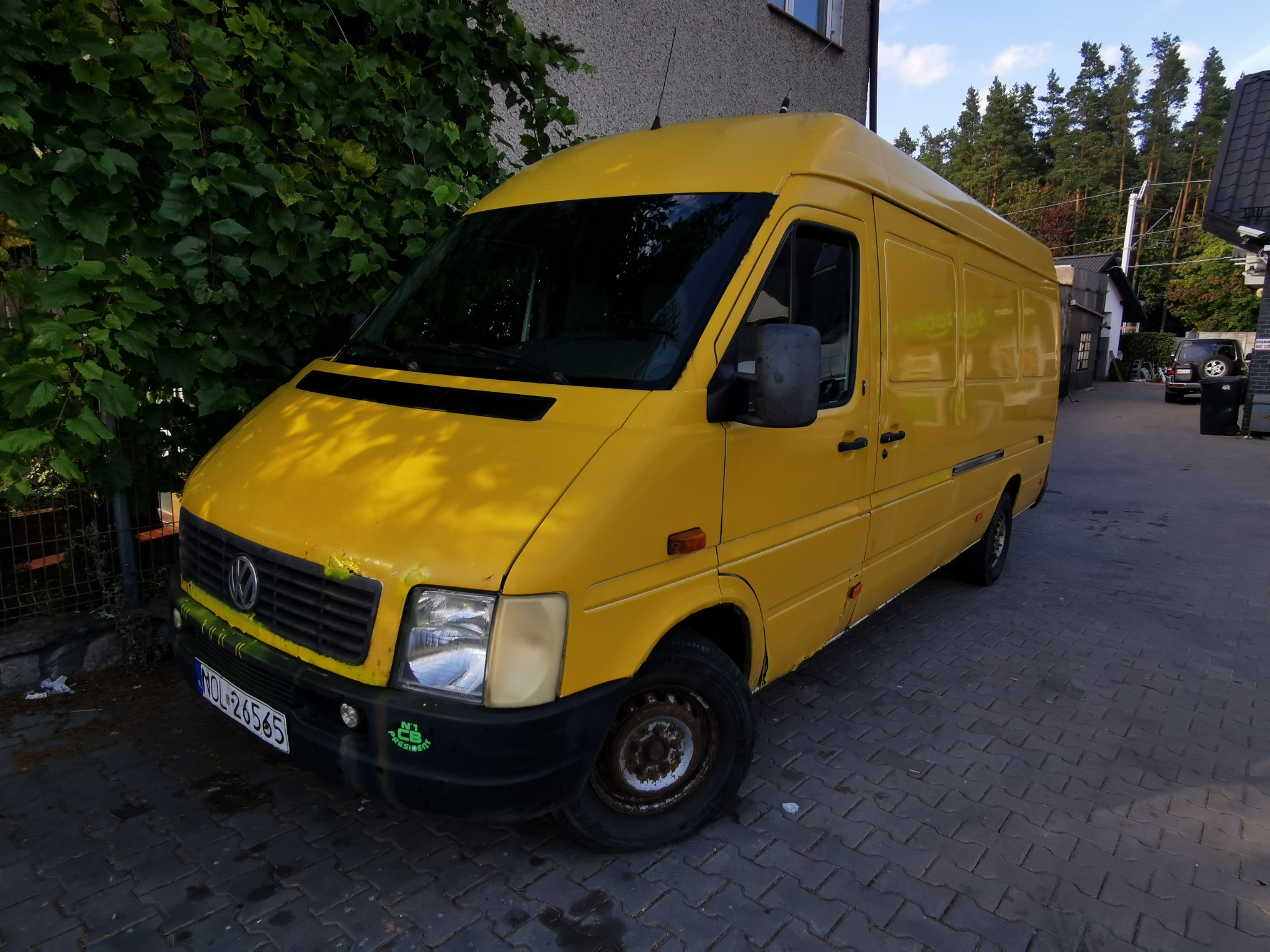 Żółty bus marki Volkswagen zaparkowany na brukowanej nawierzchni, z widocznymi śladami użytkowania na karoserii i zardzewiałą felgą, na tle zieleni i budynku.