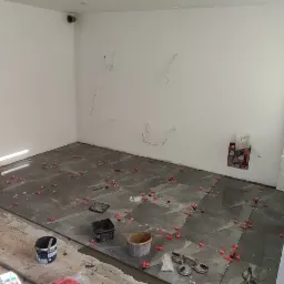 Układanie szarych płytek podłogowych w pomieszczeniu z białymi ścianami, widoczne narzędzia glazurnicze, krzyżyki dystansowe i przyssawki do płytek.